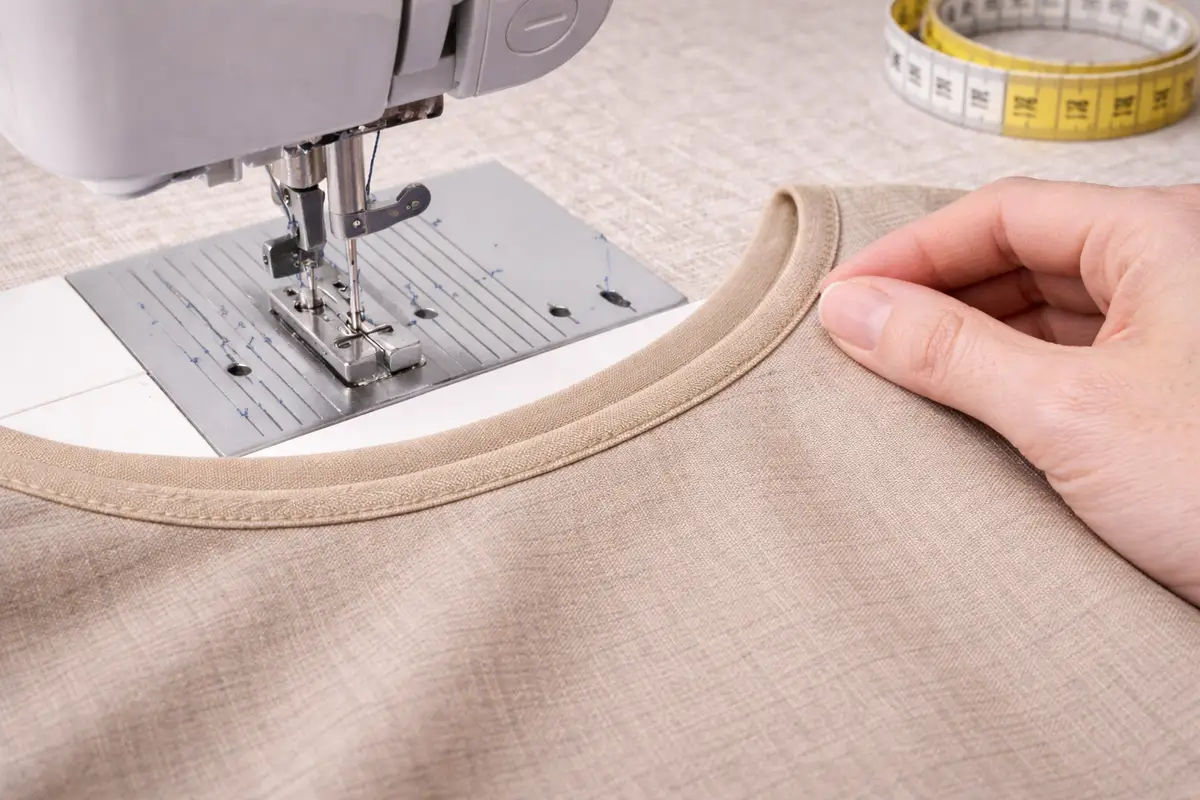Machine à coudre réalisant une surpiqûre fine sur une encolure beige pour maintenir le biais en place